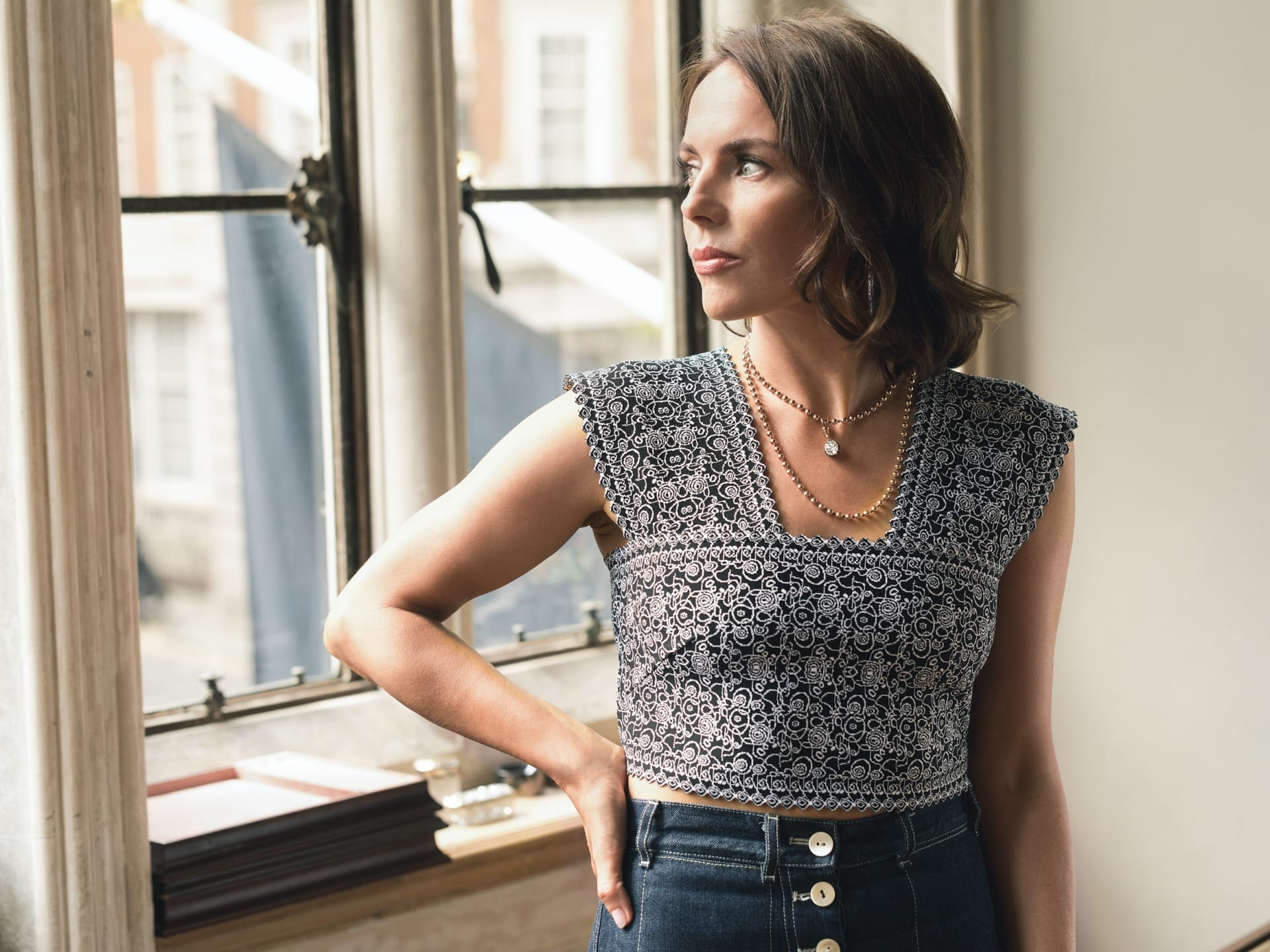 Woman in patterned sleeveless top and high-waisted jeans, standing by window with thoughtful expression.