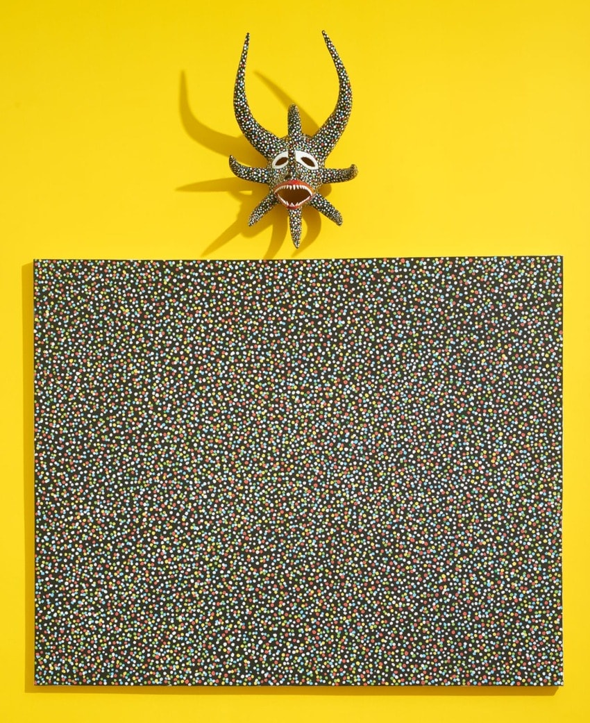 A colorful tribal mask hanging on a bright yellow wall above a black and white dotted canvas painting.