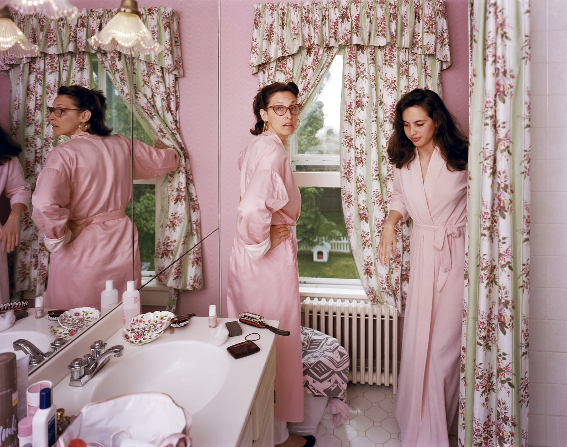 Two women in pink robes in a vintage-style pink bathroom with floral curtains, standing near a sink and a mirrored wall.