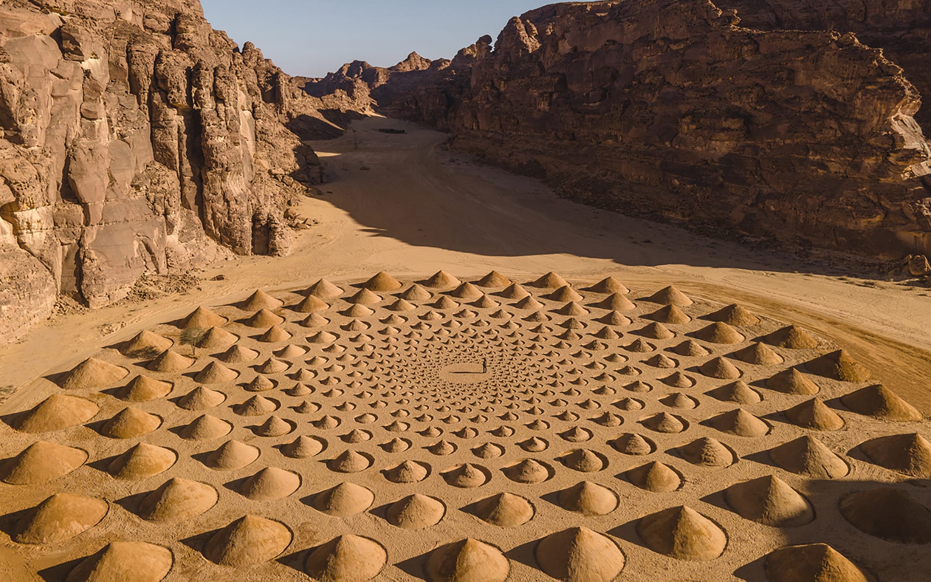 Aerial view of a stunning desert landscape with circular sand formation surrounded by rocky cliffs under a clear blue sky.