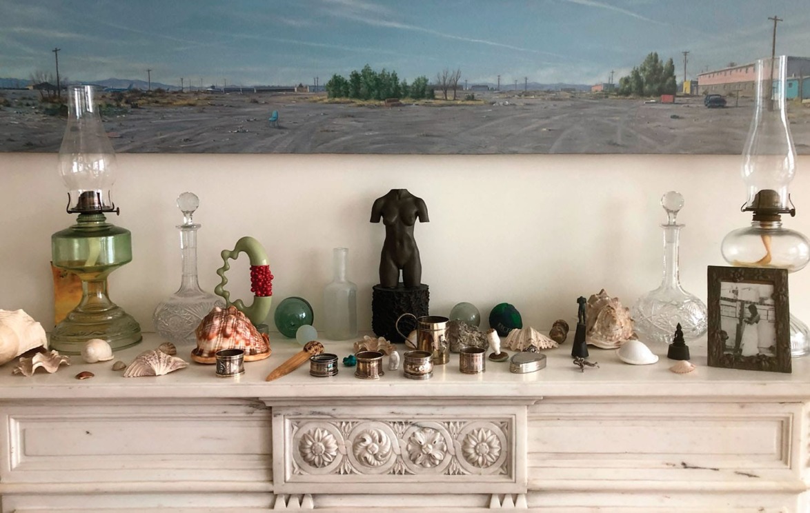 An eclectic collection of shells, lamps, and art objects arranged on a marble mantel above a landscape painting.