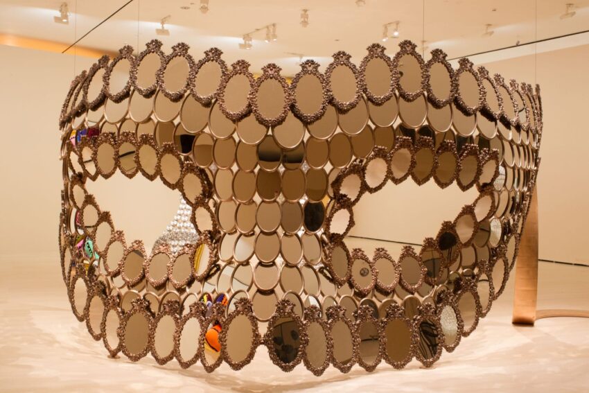 Large mirrored mask made of circular pieces on display in a brightly lit modern art gallery setting.