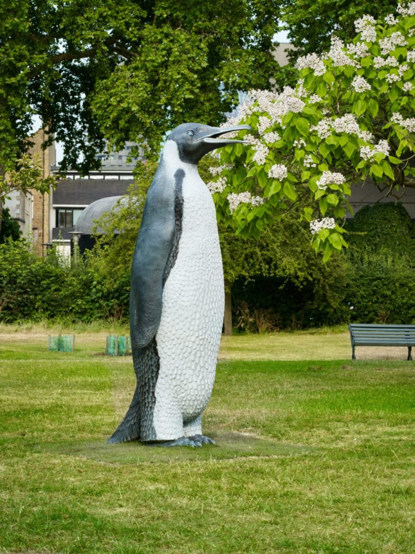 Statue of a penguin standing on grass near a tree with blooming flowers in an outdoor park setting.