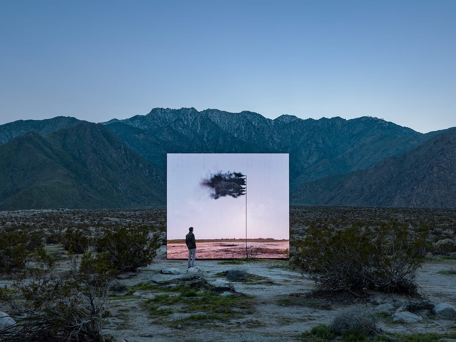 Person standing before a large reflective screen displaying a black cloud in a mountain desert landscape.