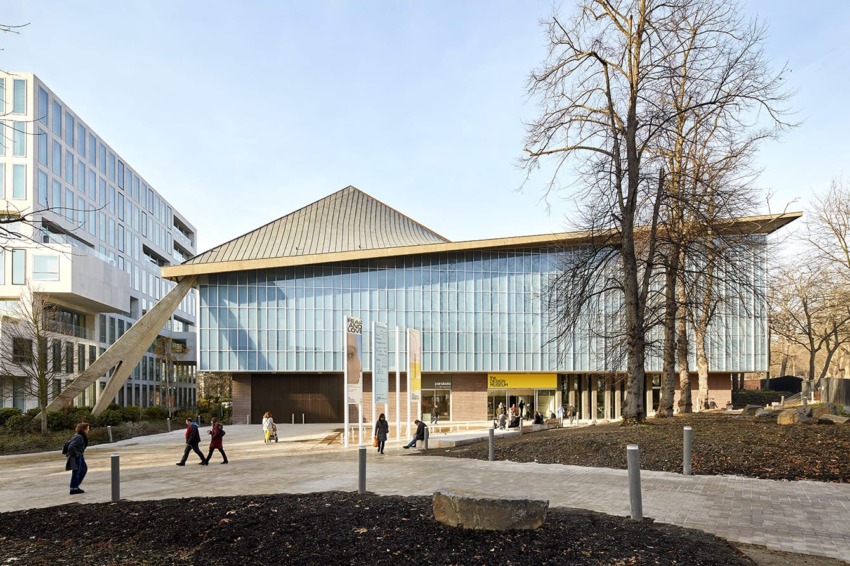 Modern building with a slanted roof, glass facade, and people walking nearby, surrounded by trees and a paved walkway.