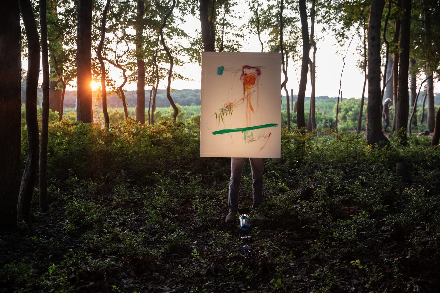 Person standing in a forest holding an abstract painting with vibrant colors, sunset visible in the background.