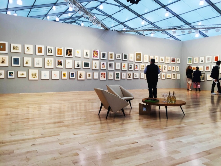 Art gallery with framed pictures on gray walls, wooden floor, modern chairs, and visitors observing the artwork.