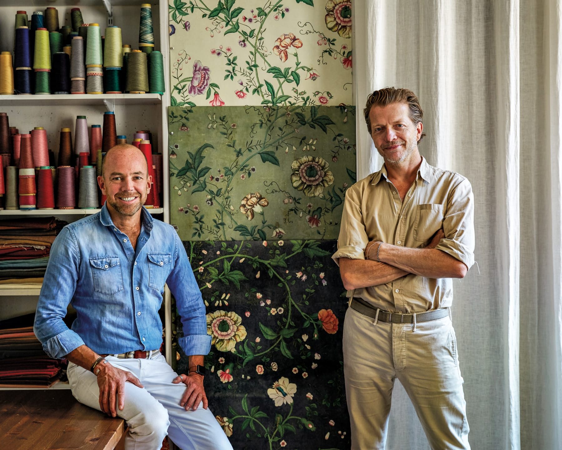 Two men in a room with spools of thread and floral tapestry designs on the wall, one seated and one standing with arms crossed.