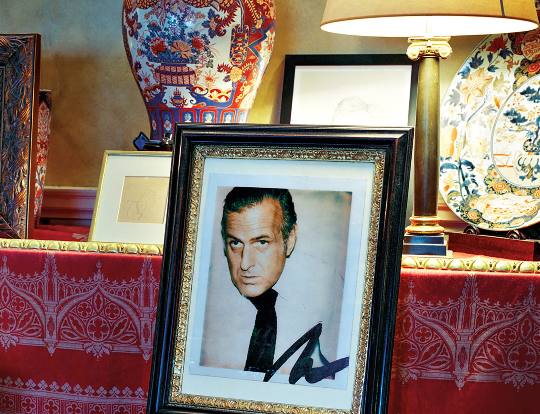 Framed photo of a man with a serious expression displayed among decorative vases and artworks on a patterned tablecloth.