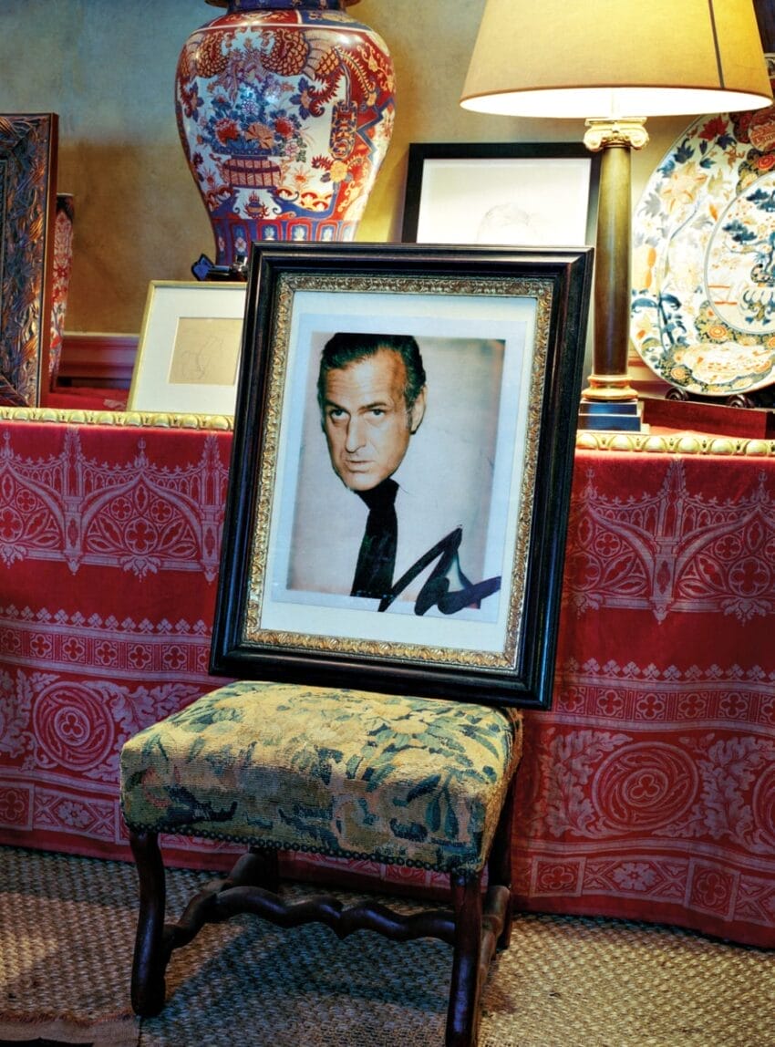 Framed black-and-white portrait on an antique chair next to a lamp and decorative vase in a richly decorated room.