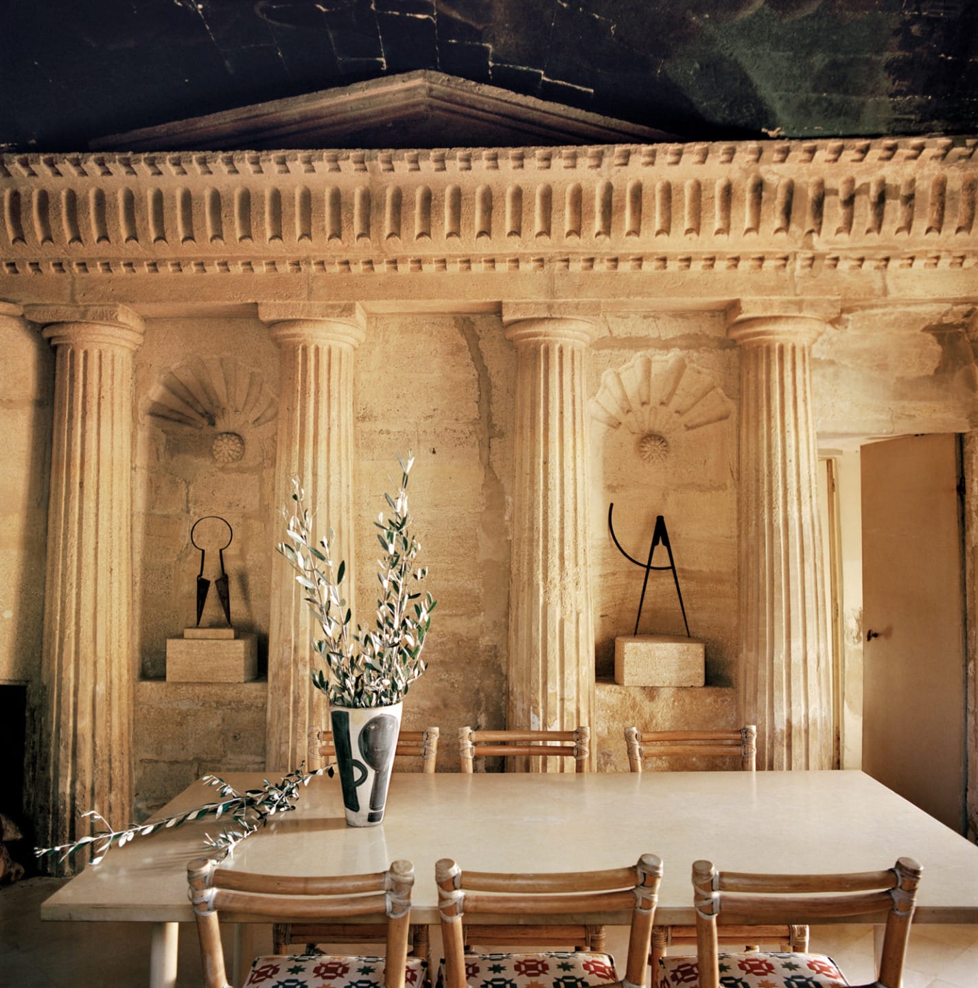 Ancient stone dining room with four pillars, a table with a vase of branches, and carved wall decorations.