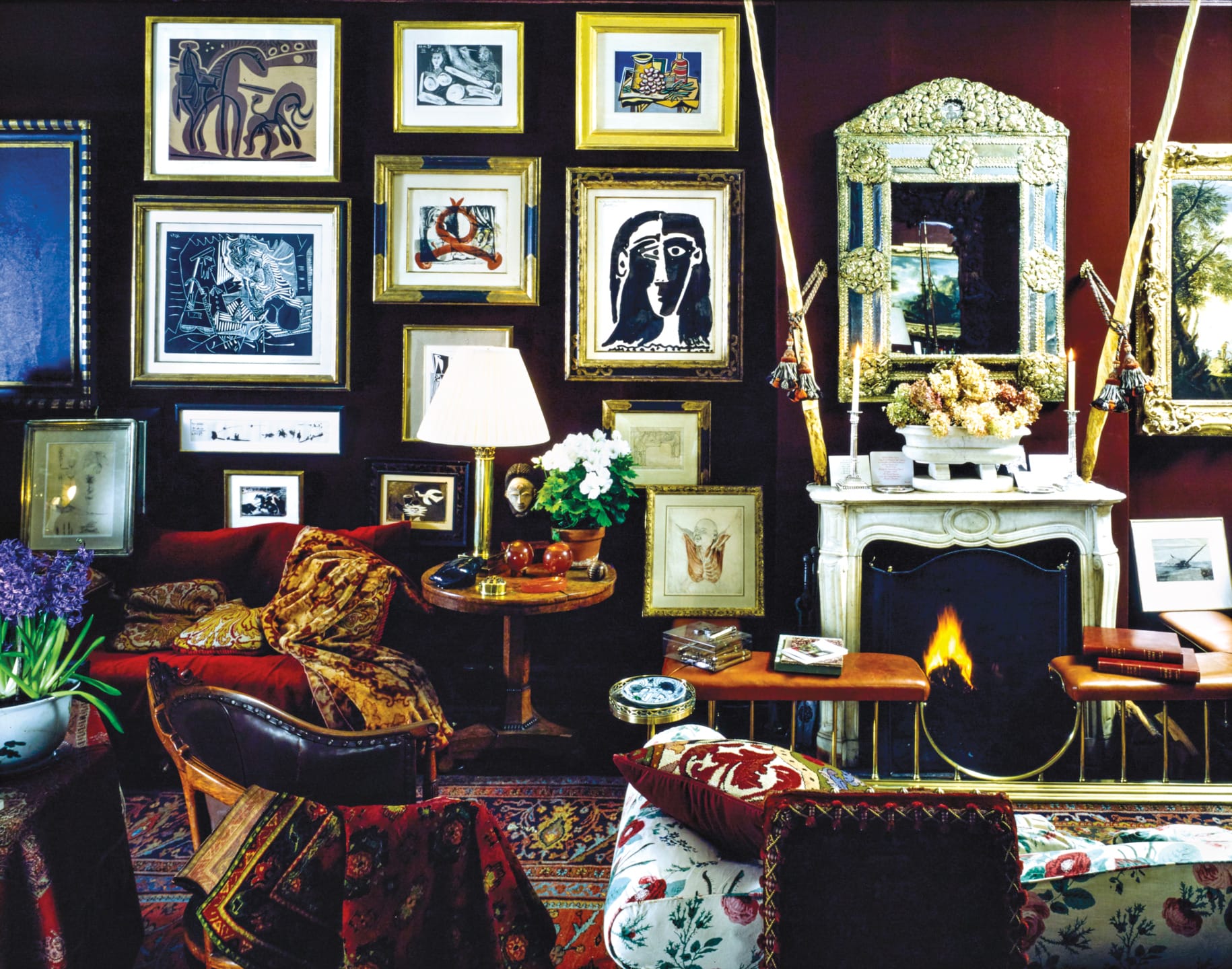Eclectic living room with art-covered walls, a floral sofa, ornate fireplace, and cozy seating around a lamp-lit table.