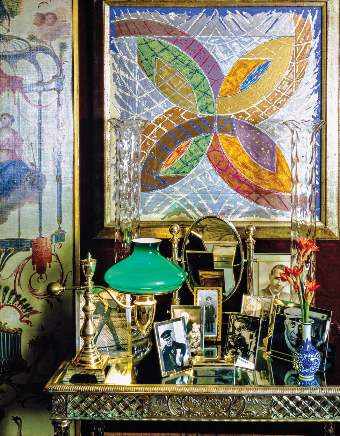 Eclectic room decor with colorful geometric painting, antique lamps, framed photos, and a vibrant flower arrangement.