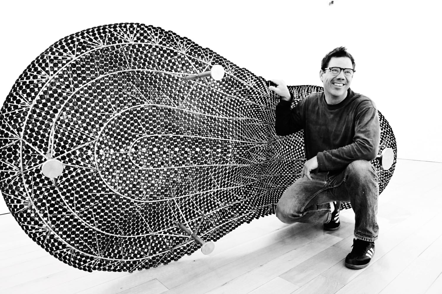 Man smiling next to large textured abstract art piece on wooden floor in a modern gallery setting.