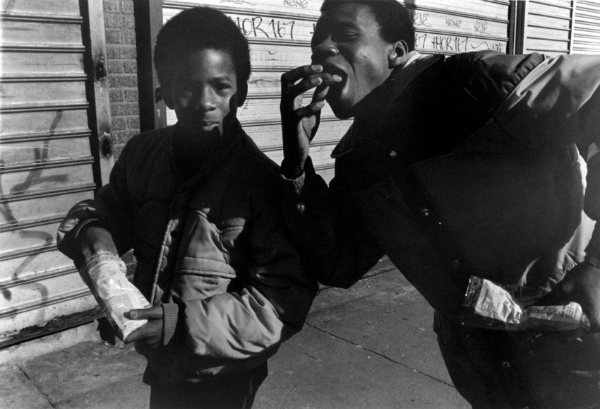 Two people standing in an urban setting, one holding a bag, both wearing jackets, with graffiti on the shuttered background.
