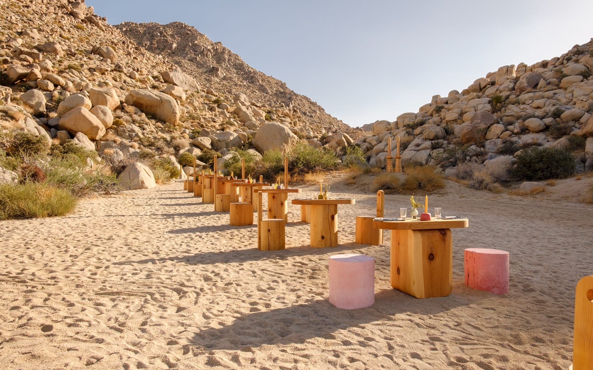 Outdoor dining setup with wooden tables and stools in a desert landscape with rocky mountains and clear sky in the background.