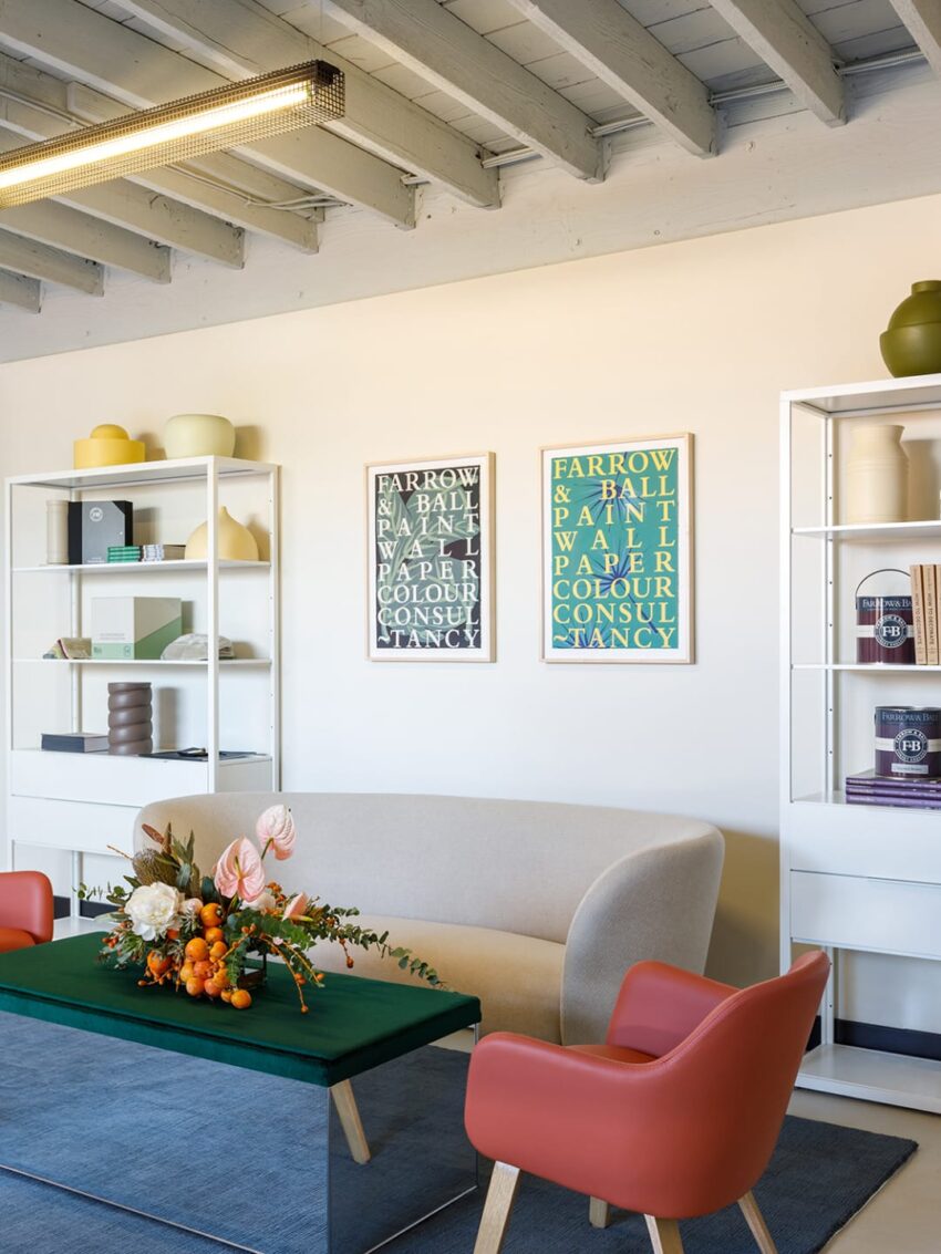 Modern office lounge with a beige sofa, orange chairs, floral centerpiece, colorful posters, and white bookshelves.