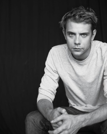 Man in a light sweater and jeans sitting on a stool against a dark background, hands clasped, looking at the camera.