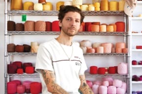 Person sitting in front of shelves filled with colorful yarns, wearing a white t-shirt with tattoos visible on their arms.