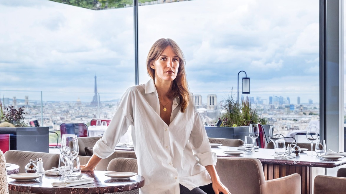 Woman seated in a modern restaurant with a panoramic view of Paris, featuring the Eiffel Tower in the background.