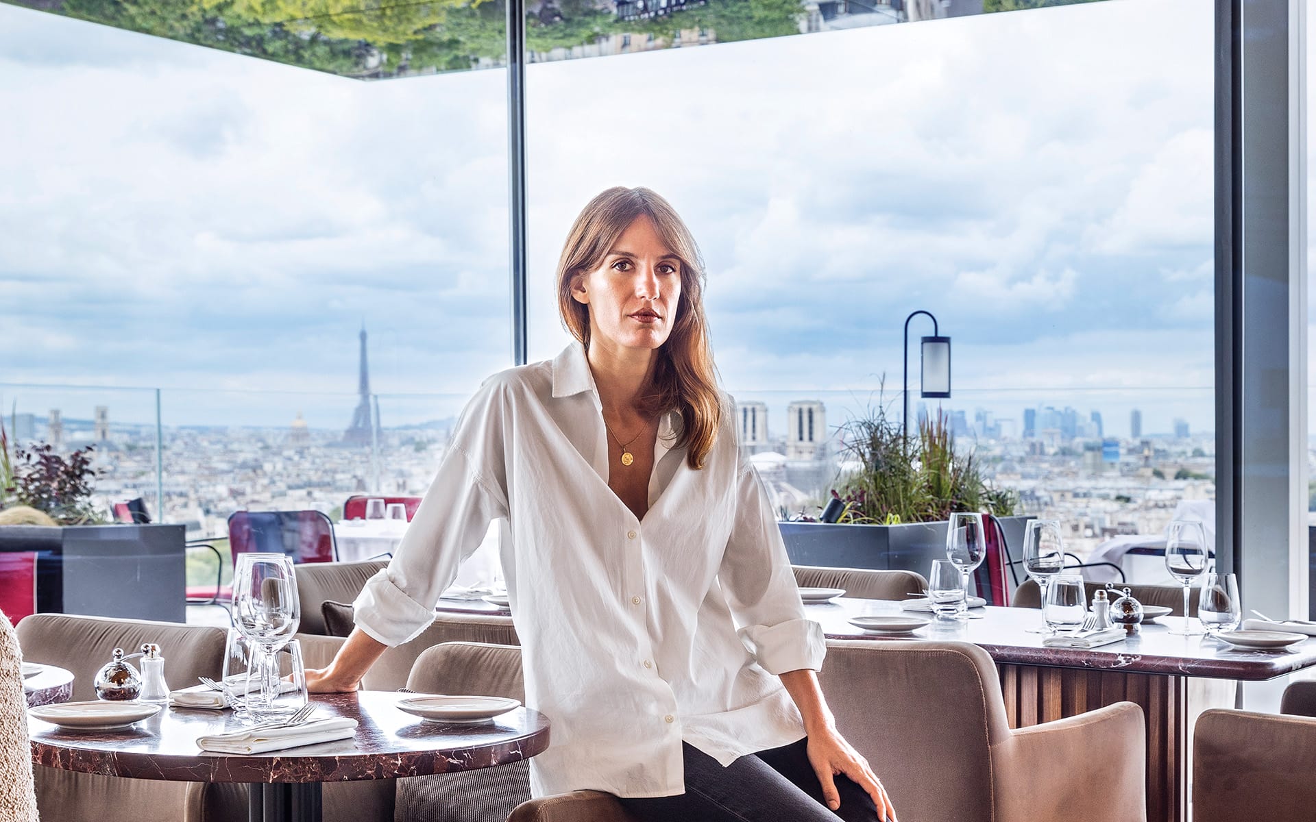 Woman seated in a modern restaurant with a panoramic view of Paris, featuring the Eiffel Tower in the background.