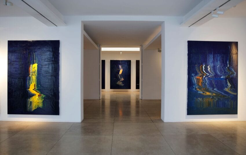 Art gallery with abstract blue and yellow paintings on white walls, polished concrete floor, and soft lighting.