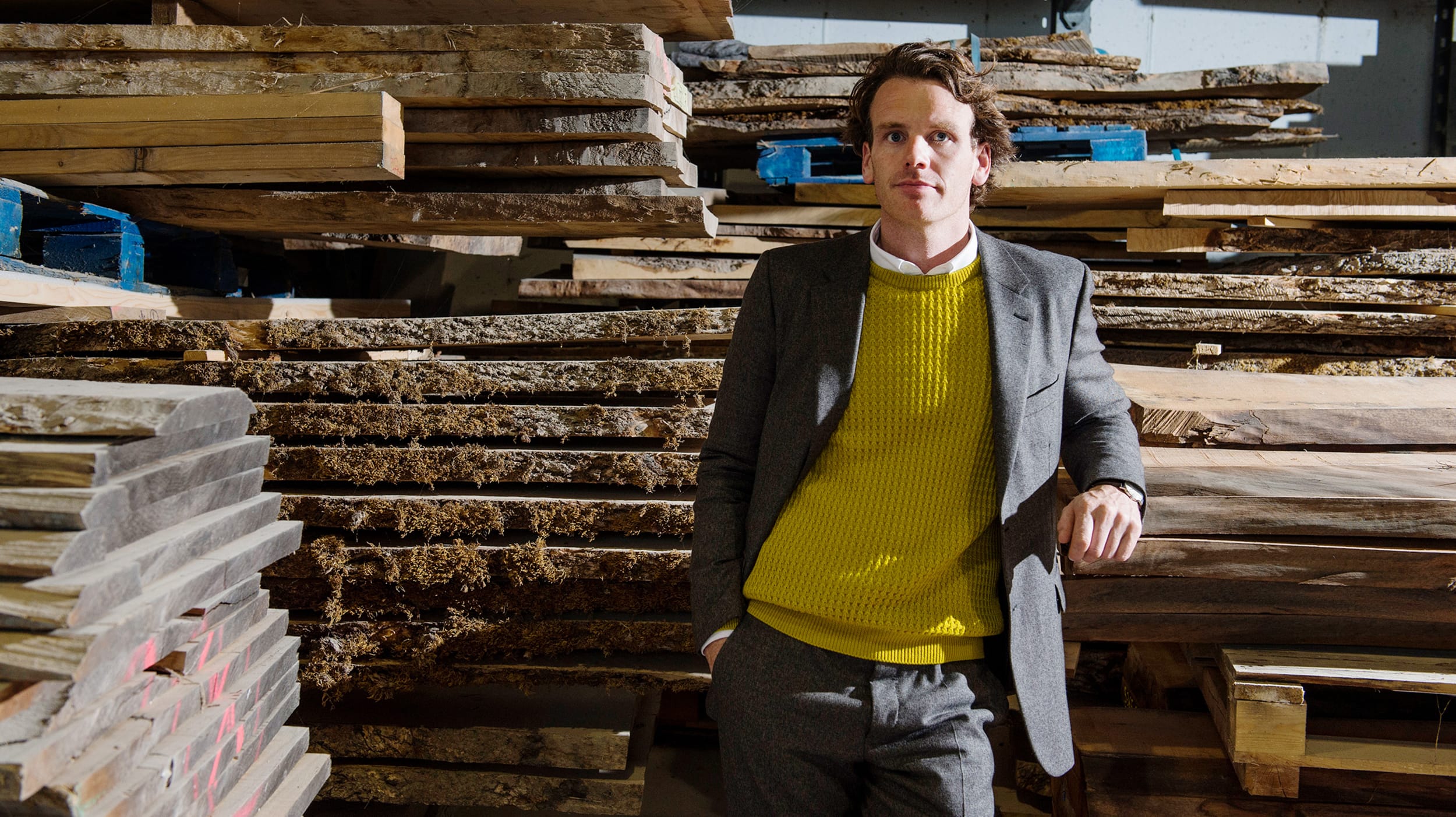Person in a yellow sweater and grey suit stands next to stacked wooden planks in a warehouse-like setting.