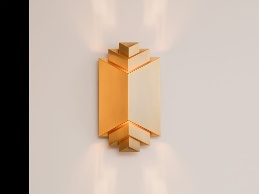 Modern geometric wall sconce with a warm gold finish against a neutral wall, creating ambient light.