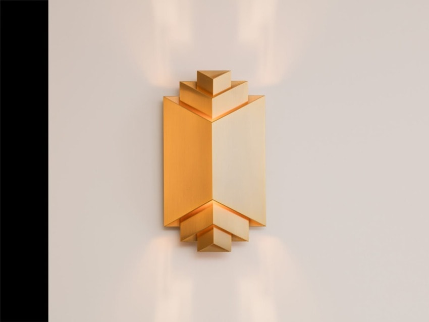 Modern geometric wall sconce with a warm gold finish against a neutral wall, creating ambient light.