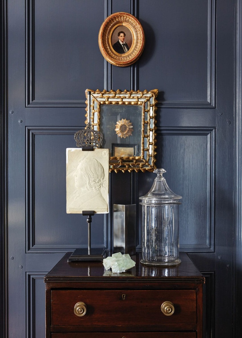 Dark wood-paneled wall with framed art, bust sculpture on stand, and a glass jar atop a wooden dresser.