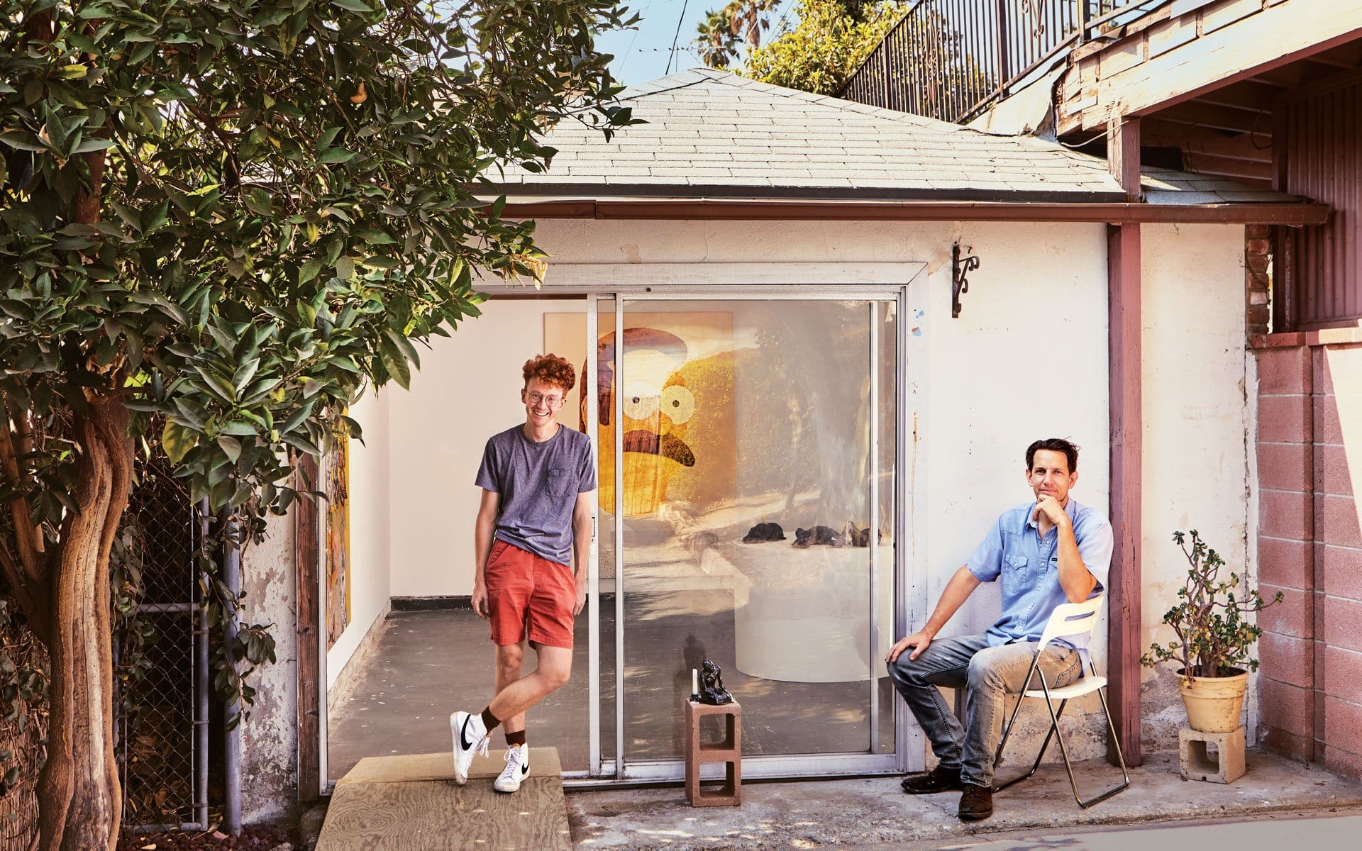 Two men sitting outside a small art studio with a colorful painting visible through sliding glass doors.