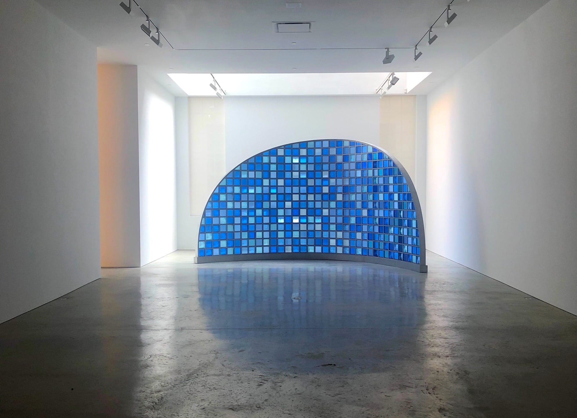 Art installation in a gallery with a curved wall featuring a mosaic of blue tiles and illuminated by overhead lights.