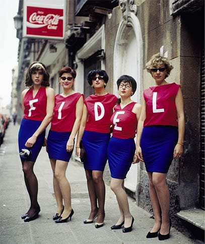 Five people wearing red tops and blue skirts, standing in a row, each top displaying a letter spelling "FIDEL" on a city street.