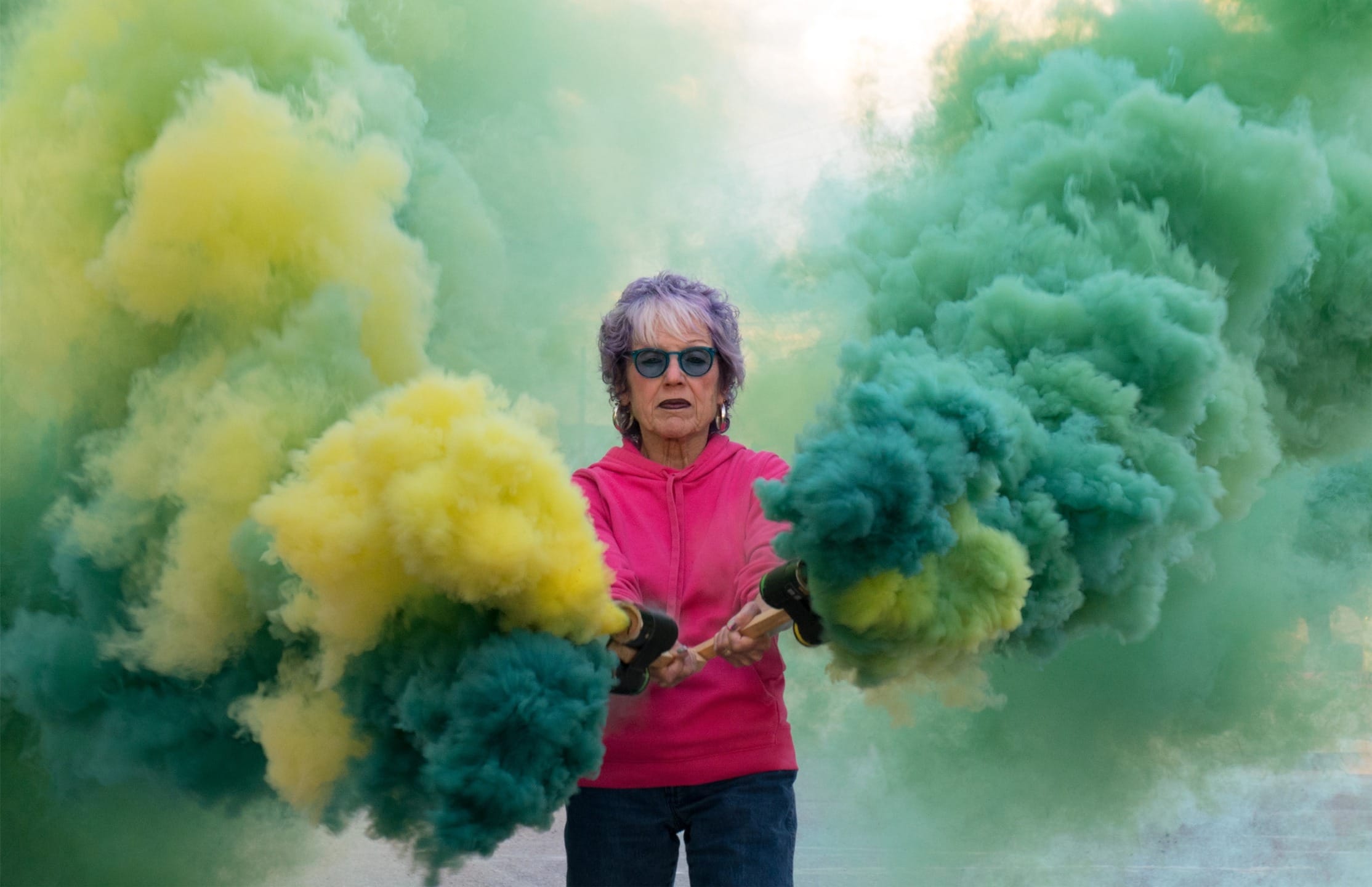 Person in a pink hoodie surrounded by colorful yellow and green smoke outdoors.