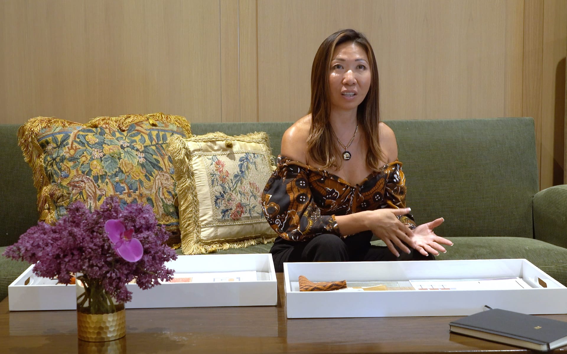 Woman sitting on a green sofa, explaining something with hand gestures, beside decorative pillows and a vase of purple flowers.