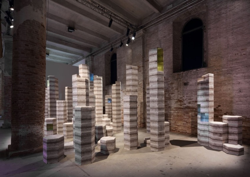 Stacks of paper resembling towers in a dimly lit industrial-style gallery space with brick walls and columns.