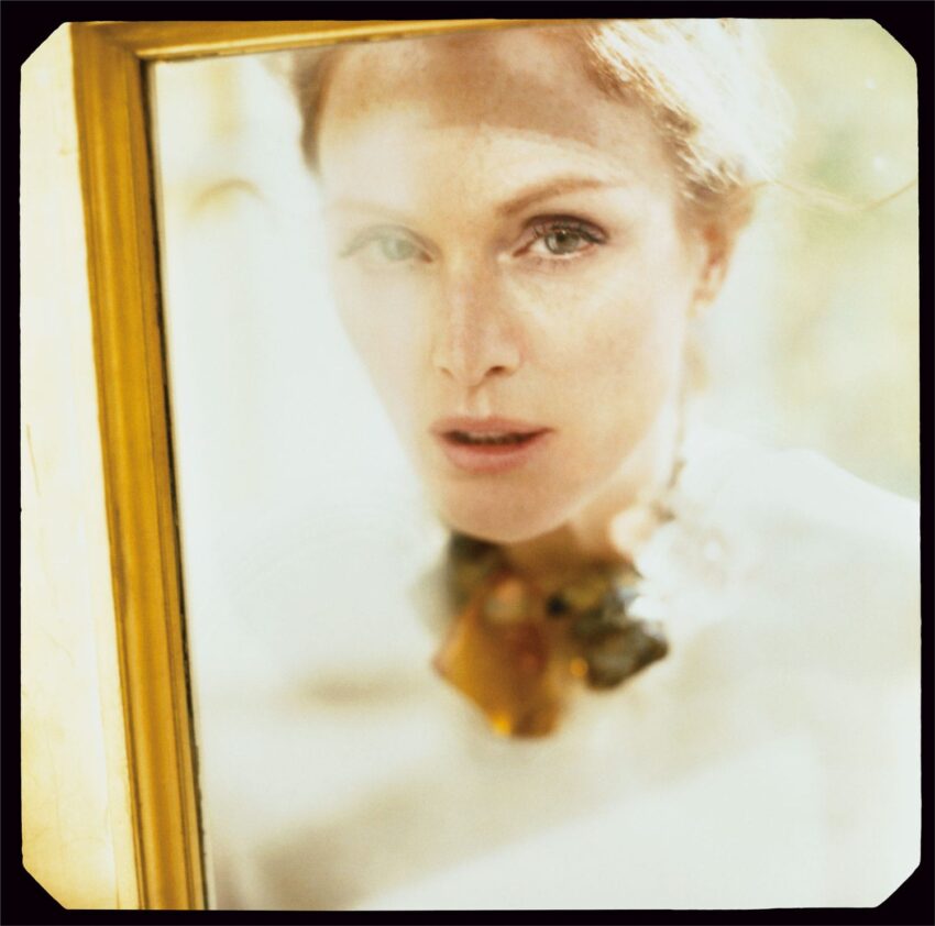 Portrait of a person with red hair looking through a glass pane, wearing a statement necklace, with soft focus effect.