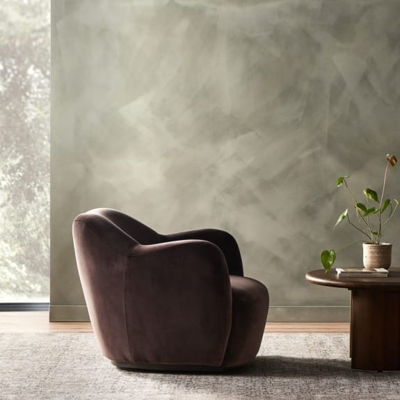 Cozy brown velvet armchair beside a wooden table with a potted plant, set against a textured green wall in a softly lit room.