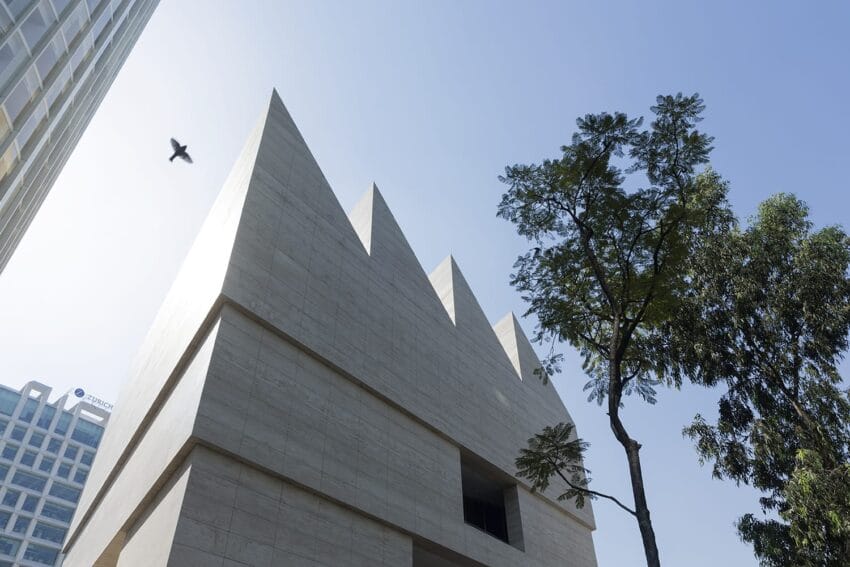 Modern triangular building with sharp edges under a clear blue sky, accompanied by a tree and a bird in flight.