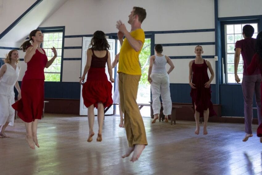 Group of people dancing in a studio with wooden floors, some wearing red outfits and one in a yellow shirt.