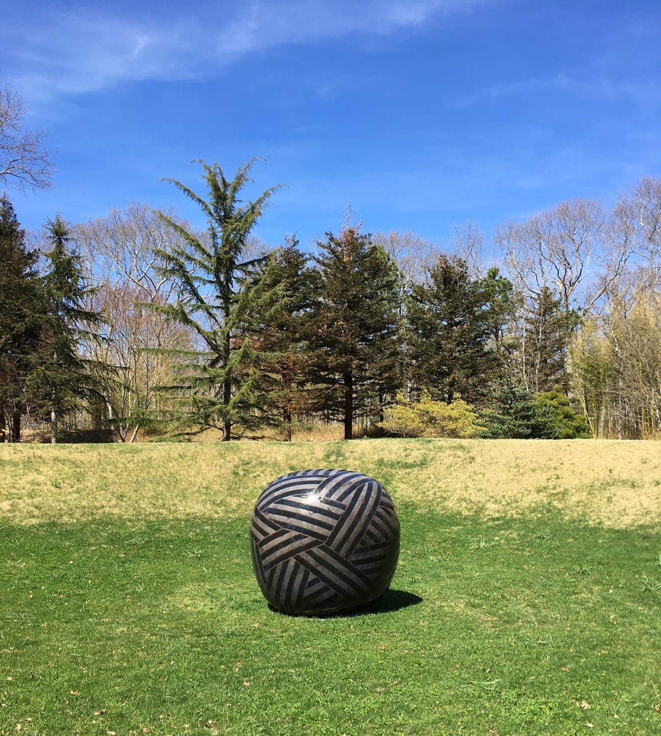 Sculpture resembling a large woven sphere on grass with trees and clear blue sky in the background.