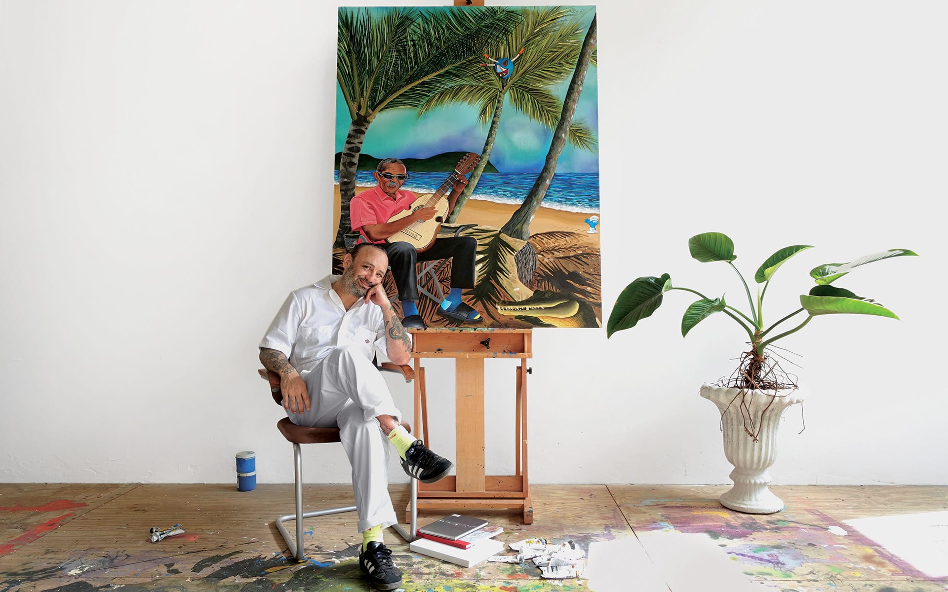 Artist sitting in a studio next to a tropical painting on an easel with art supplies on the floor.