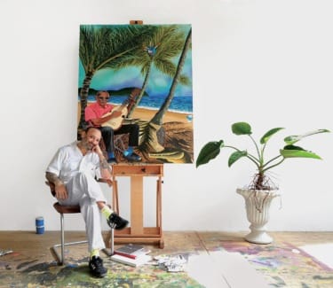 Artist sitting in a studio next to a tropical-themed painting on an easel, with a plant and paint supplies nearby.