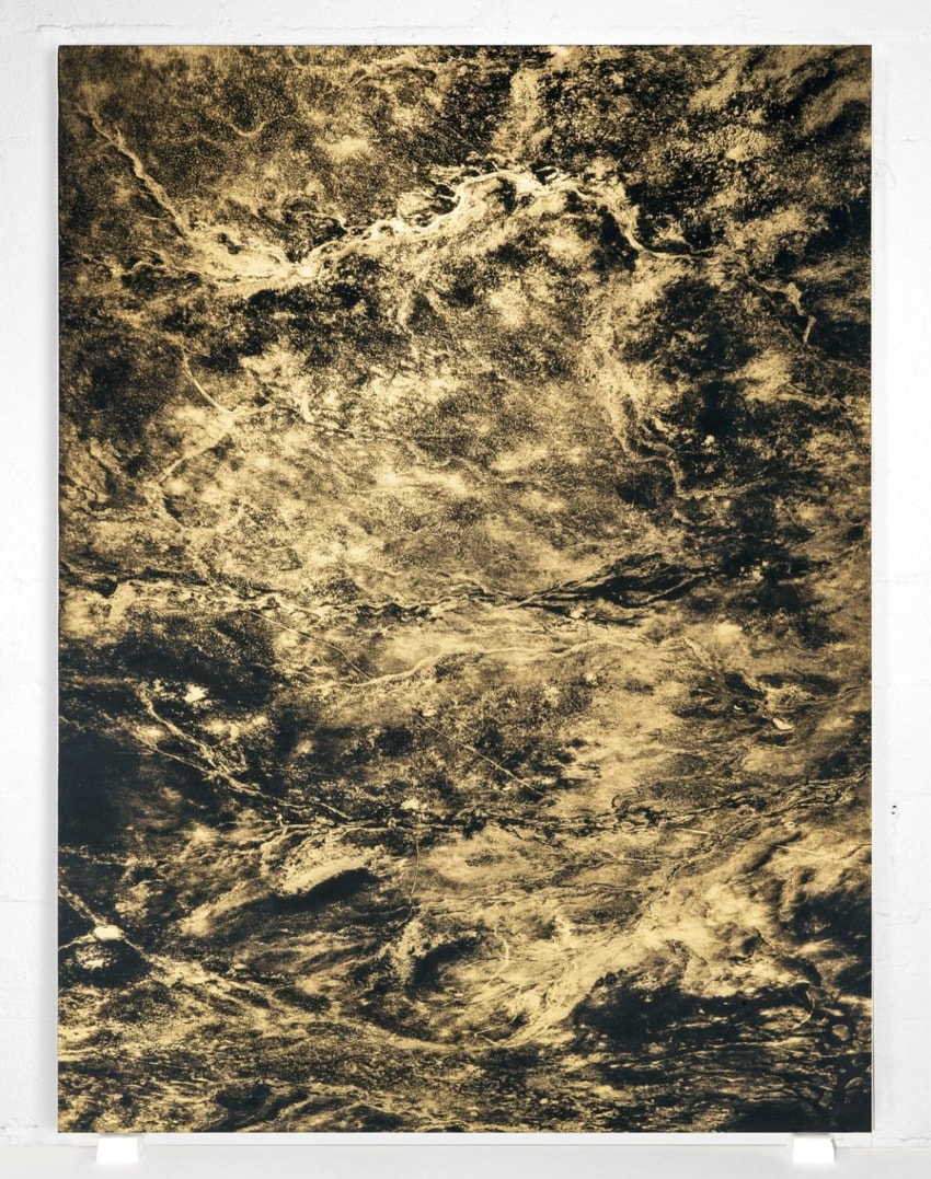 Abstract textured artwork in gold and black with swirling patterns, mounted on a light gray wall.