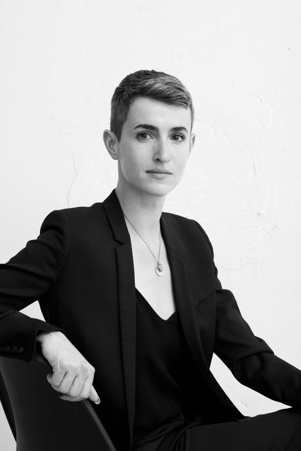 Person with short hair in a black suit sitting on a chair against a white textured background.