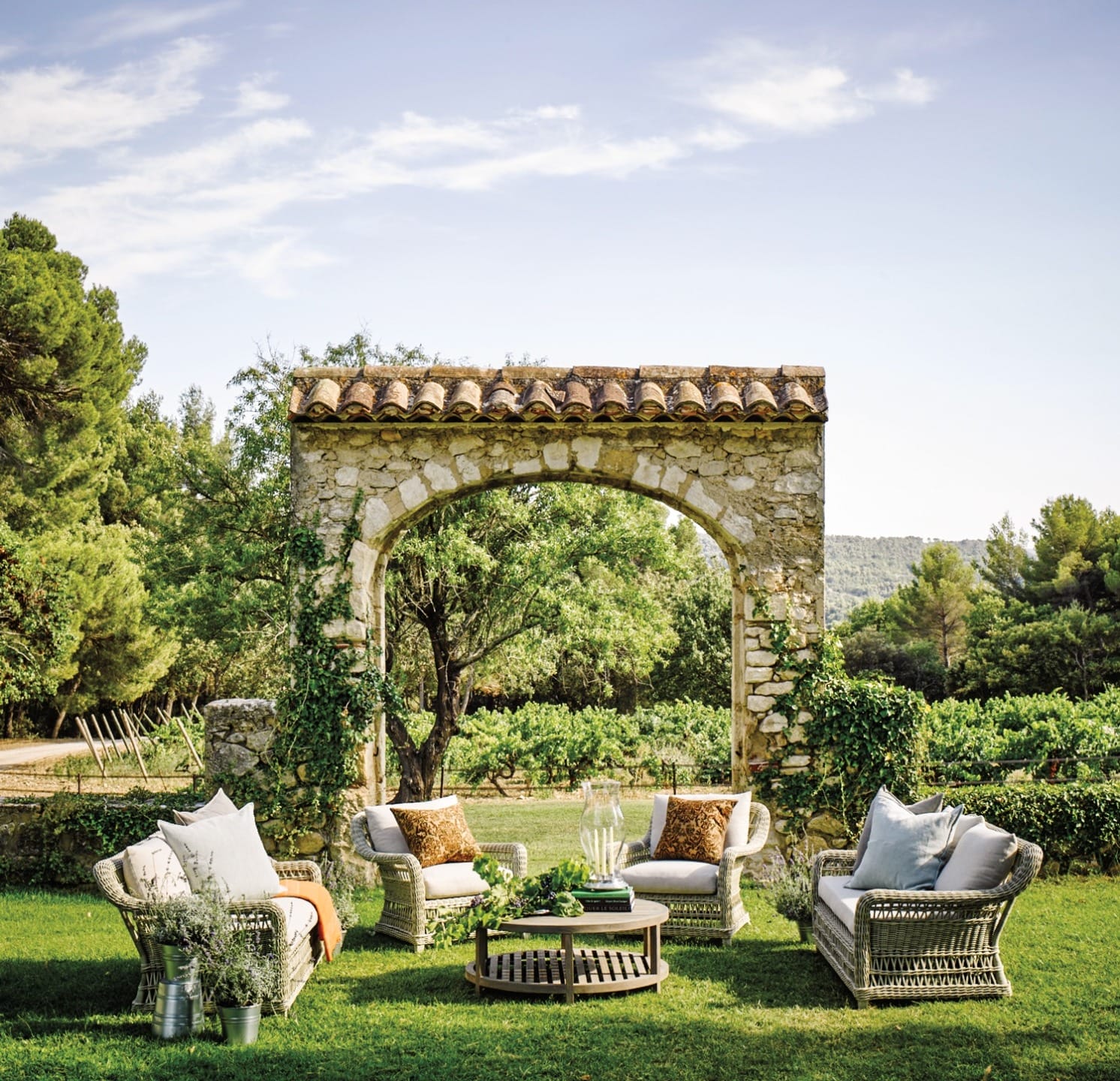Outdoor seating area with wicker furniture and stone arch surrounded by greenery under a clear blue sky.