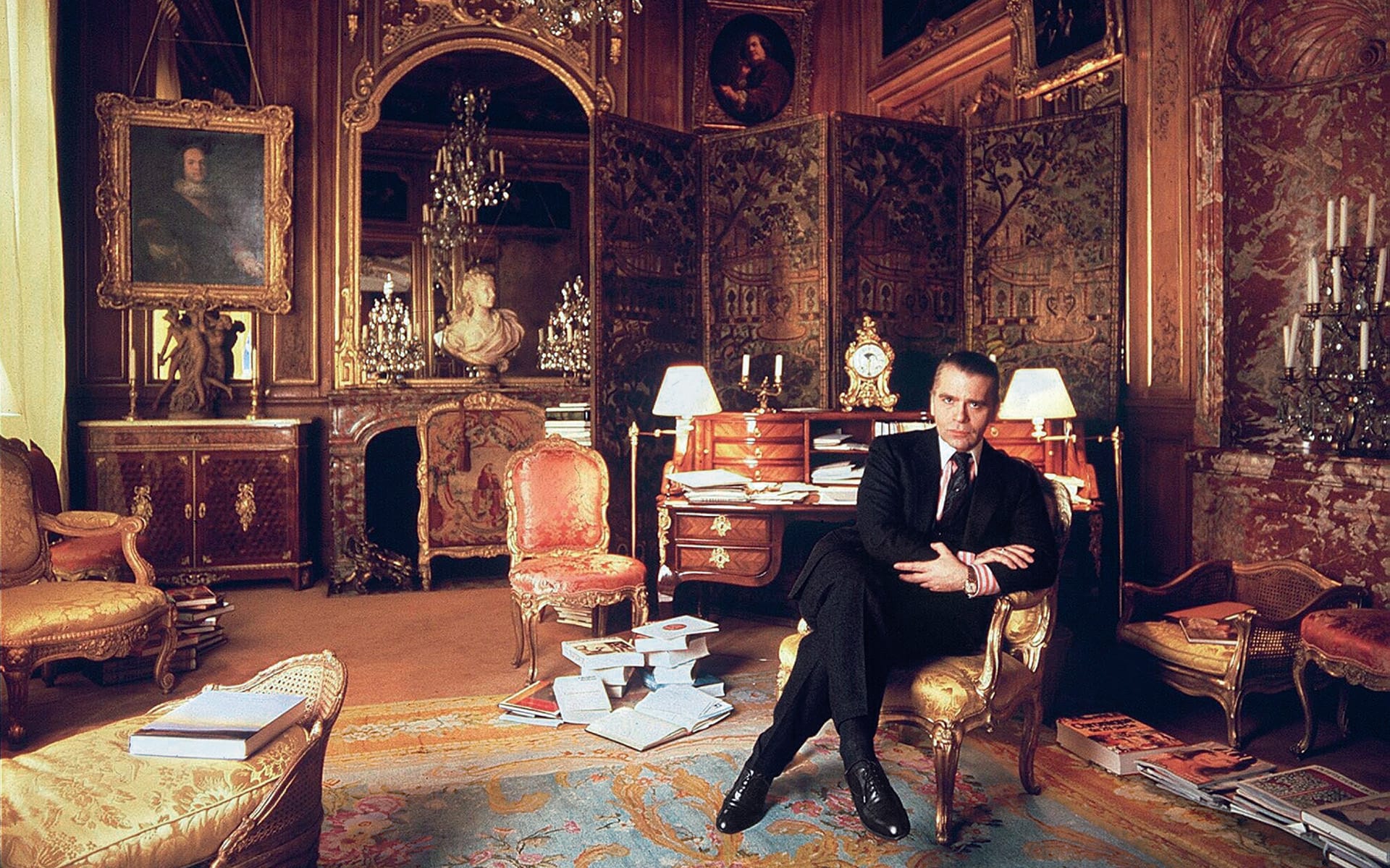 Luxurious ornate room with chandelier, antique furniture, paintings, and a person sitting among books and elegant decor.