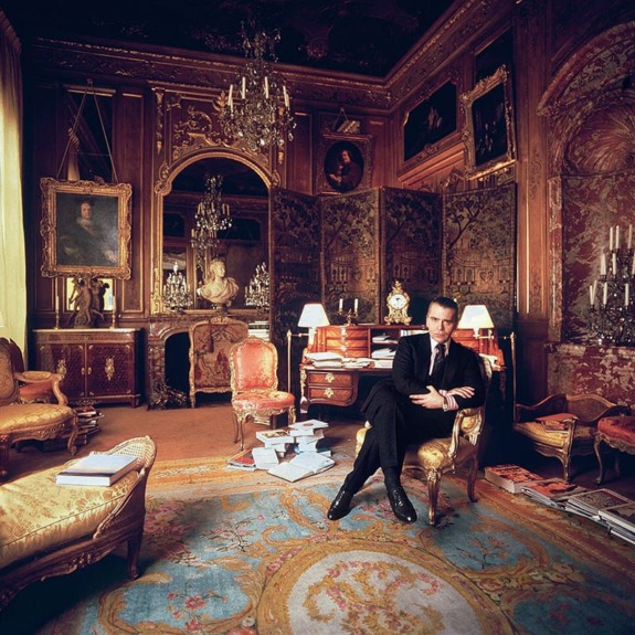 Man seated in a lavishly decorated room with ornate furniture, mirrors, and chandeliers, surrounded by books and art.