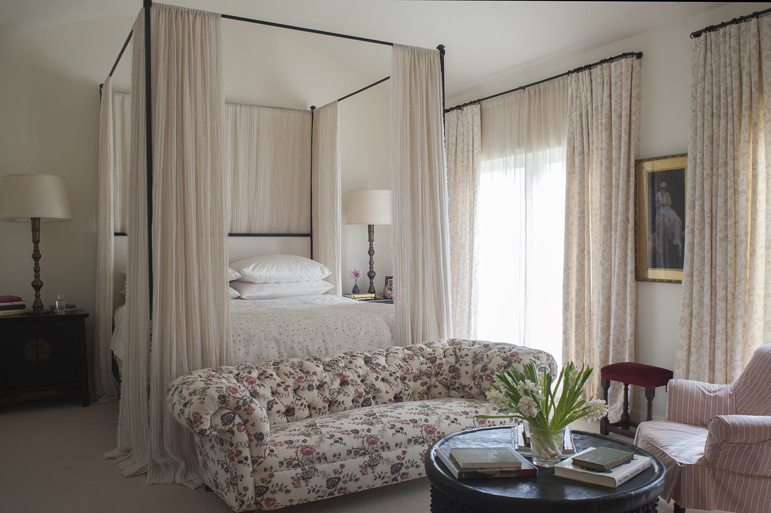 Elegant bedroom with a four-poster bed, floral sofa, books, and flowers on a black coffee table, softly lit by natural light.