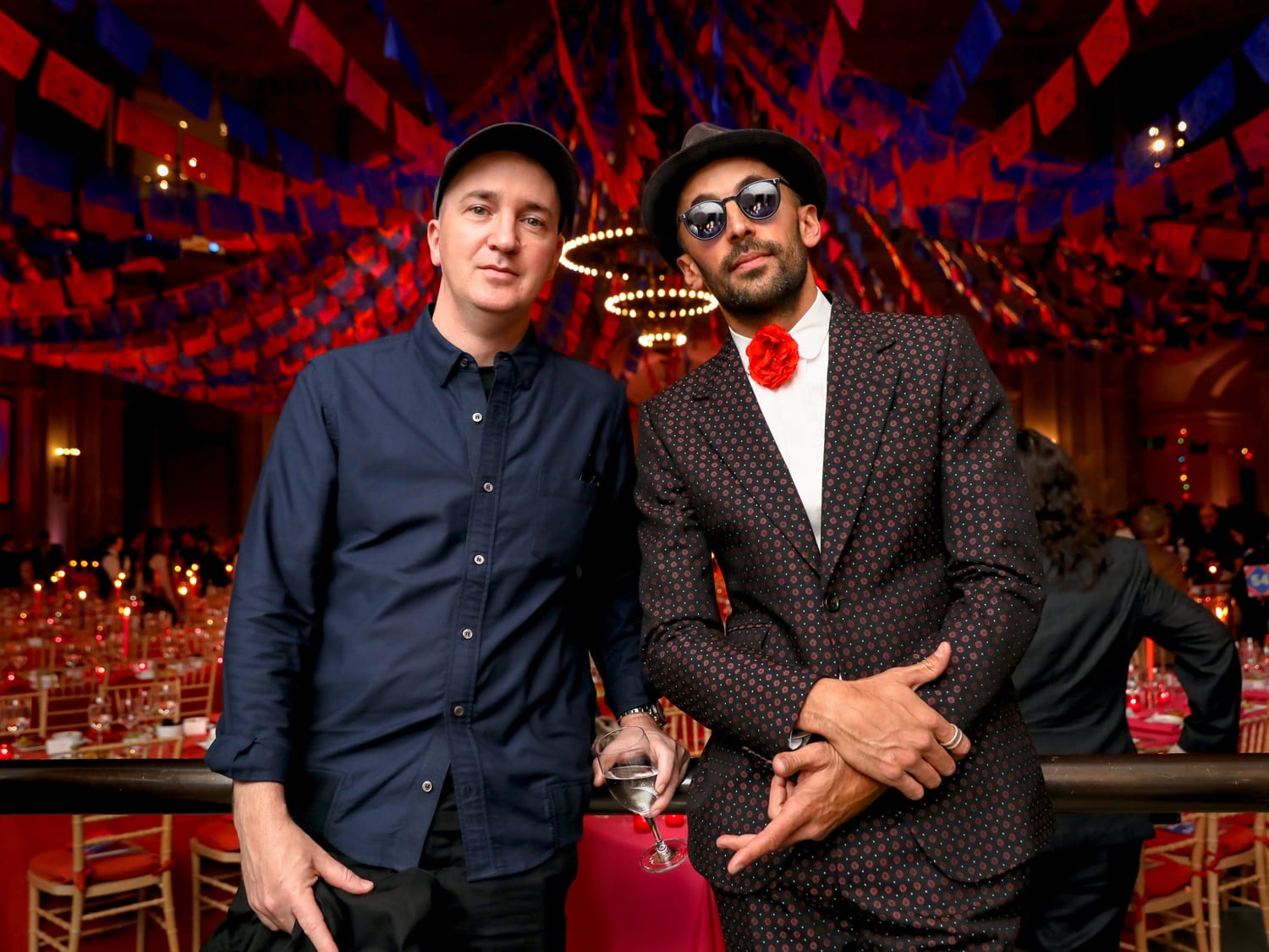 Two men posing at a festive event with colorful flags in the background. One wears a hat, the other sunglasses and a red flower.